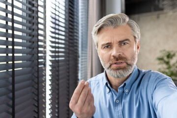 Mature man with gray hair and beard looking serious and gesturing near a window with blinds. Interior setting conveys a professional or personal communication.