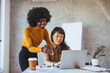 © Dragana Gordic - An Asian and a Black businesswoman engage in a collaborative effort, reviewing data on a laptop in a modern office setting, professional attire reflecting their entrepreneurial roles.