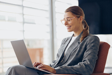 Wall Mural - Stylish female freelancer in eyeglasses working on laptop while sitting in modern coworking 