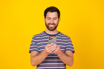 Poster - Photo of positive optimistic man with beard dressed striped t-shirt look at smartphone read sms isolated on yellow color background