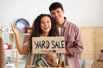 Canvas Print - Young couple holding cardboard with text YARD SALE in room of unwanted stuff