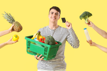 Wall Mural - Young man holding full shopping basket with credit card and food on yellow background