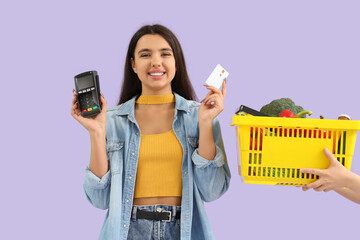 Poster - Young woman holding payment terminal with credit card and full shopping basket on lilac background