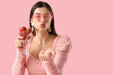 Wall Mural - Beautiful young woman holding glass with fresh strawberries and blowing kiss on pink background