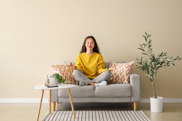 Wall Mural - Young Asian woman meditating on grey sofa near beige wall