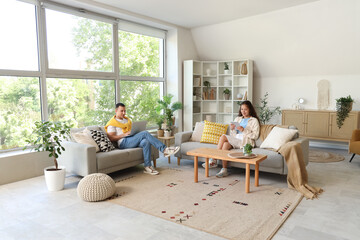 Wall Mural - Young couple using modern gadgets on sofas at home