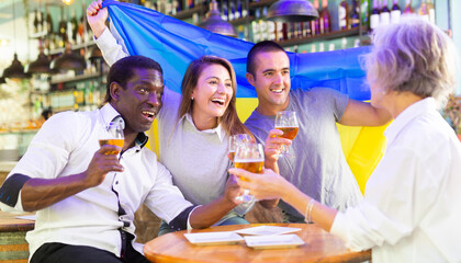 Wall Mural - Excited multiracial soccer fans with flag of Ukraine celebrating victory of team with pint of beer in the pub