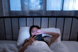 © MergeIdea - Asian young woman lying in bed at night holding a smartphone and covering her eye from the screen brightness of blue light unhealthy lifestyle before sleep