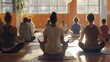 © Emiliia - A group of people meditates in a yoga studio, with sunlight streaming through large windows.