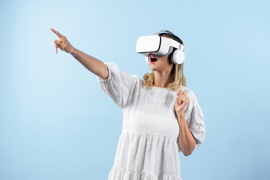 Caucasian girl in pajamas enter in metaverse while point at blue background. Excited woman in white dress enjoy connecting in visual reality world program. Technology innovation concept. Contraption.