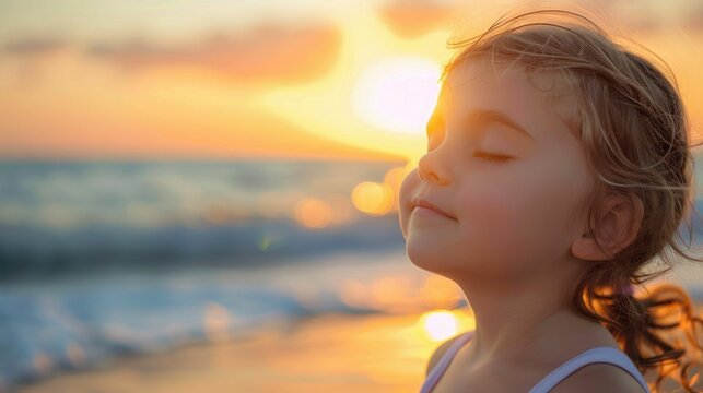 A happy little girl sits on the beach with her eyes closed, feeling the warmth of the sunlight on her face. Her nose wrinkles in a smile as she listens to the water and feels the breeze in her hair