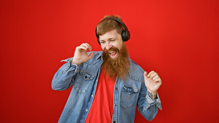 Confident young redhead guy loving his music, dancing and having fun, over an isolated red background while listening in casual attire, oozing positivity and happiness