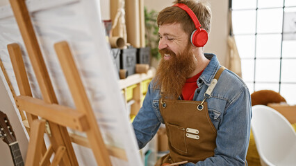 Poster - Handsome, young, redhead bearded man, joyfully engrossed in a painting lesson, listening to favorite songs, while standing at an easel drawing in an art studio, wearing an apron, smiling.