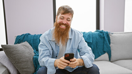 Wall Mural - Handsome young redhead man cheerfully using his smartphone while sitting on the sofa in the living room of his home, spreading joy and positive vibes through texting and the online world