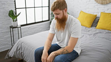 Sticker - Handsome, young irish redhead man in a relaxed morning lifestyle, sitting on his bedroom's bed, looking down with a serious expression of concentration on his face indoors