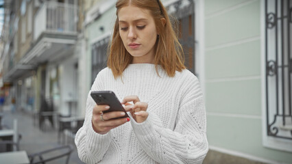 Wall Mural - A young woman in a white sweater looks at her phone on a quiet city street.