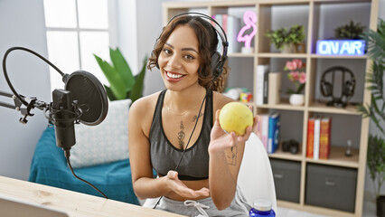 A smiling young woman with headphones holding an apple in a modern radio studio, representing health and lifestyle podcasting.