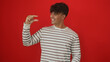 © Krakenimages.com - A young hispanic man wearing a striped shirt, smiling and gesturing with his hand as if holding something small, stands against a vibrant red background.