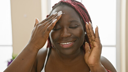 Sticker - A smiling woman with braids applies cream to her face indoors, portraying a moment of beauty routine.