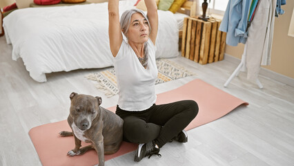 Poster - Sporty grey-haired middle-aged woman with pet dog, training yoga in serene bedroom ambiance, sitting on floor near bed for healthy workout regime