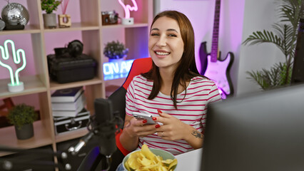 Wall Mural - A smiling young woman using smartphone in her colorful gaming room filled with neon lights, guitar, and snacks.