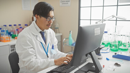 Young asian male scientist working on a computer in a laboratory setting with various equipment in the background.