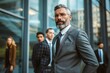© anatolir - Poised male executive with a beard stands before a modern office building, with a team in the background