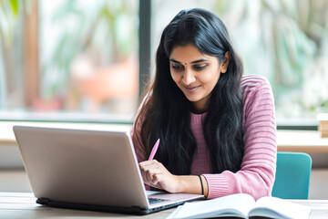 Young adult indian student woman taking notes while using laptop computer at home. Millennial ethnic female learning online listening virtual video call. Business and education concept