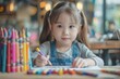 © easybanana - The little girl is sitting at a table and drawing with colored pencils. She is wearing a denim jumper and has her hair in pigtails. She is looking at the camera with a serious expression.