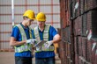 © BESTIMAGE - Two warehouse workers wearing hard hats and safety vests are looking at a tablet while standing in front of a large stack of metal beams.