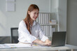 © amnaj - Smiling young woman working on a laptop in a bright office environment. Focused on her task, demonstrating productivity and enthusiasm at work.