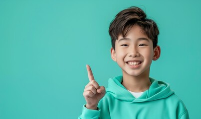 Wall Mural - Smiling asian teenager boy in a mint sweatshirt smiling with healthy teeth and pointing index finger up at copy space expressing wow emotion on a mint background