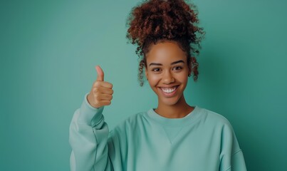Wall Mural - Young curly african american girl in a sweatshirt smiling with healthy teeth showing thumb up at copy space expressing wow emotion standing isolated on mint background