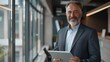 © Mahemud - Smiling middle-aged businessman, CEO, or investor using a tablet computer, mature older executive looking at the camera while promoting finance trading work in an office.