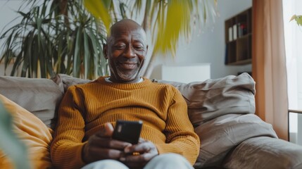 A man in a yellow sweater is sitting on a couch and looking at his cell phone