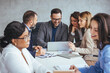 © Dragana Gordic - A group of professional men and women of different ethnicities actively participate in a business meeting, discussing over a laptop in a modern office setting.