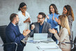 © Dragana Gordic - A group of diverse business professionals celebrate success, clapping for a smiling male colleague seated with a laptop in a modern office.