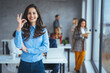 © Dragana Gordic - A smiling Caucasian businesswoman gives an OK sign, dressed professionally, indicating success. Colleagues engage in the background, highlighting a dynamic, professional setting.