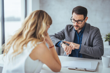 Wall Mural - A focused Caucasian businesswoman and businessman engage in collaboration at an office desk, discussing work with documents and a wristwatch in view.