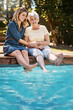 © ReadyAtTheEase/peopleimages.com - Mother, daughter and happiness by swimming pool for bonding, holding hands or relax in garden. Senior mom, woman and together in outdoor with smile for wellness, family or love and feet in water