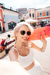 Wall Mural - Young beautiful smiling female in summer white cycling shorts and tank top clothes. Sexy carefree woman posing in street. Positive model . Cheerful and happy. Holds penny skateboard. Takes pov selfie