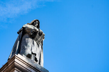 The Monument to Giordano Bruno, a bronze statue of the Italian philosopher holding a book, stands tall against a vibrant blue sky in Campo de Fiori, Rome.