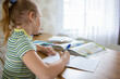 © evgeniia_1010 - Portrait of a charming blonde girl who does her homework using textbooks and a workbook. The concept of distance learning online and at home.