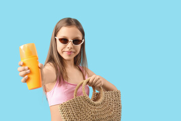 Cute little girl in swimsuit, sunglasses with wicker bag and sunscreen cream on blue background