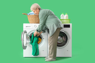 Wall Mural - Senior woman with basket of clothes putting laundry into washing machine on green background