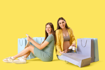 Canvas Print - Pregnant female friends with baby clothes and shopping bags on yellow background