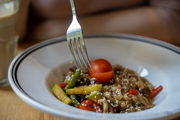 Asian pasta with sweet pepper, tomatoes, zucchini, sesame, european woman eating with a fork from a white plate in a restaurant