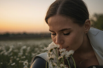 Wall Mural - A woman is sitting in a field of white flowers and smelling them. The scene is peaceful and serene, with the sun setting in the background. The woman is enjoying the moment.