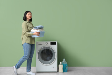 Wall Mural - Young woman with laundry walking near washing machine on green background