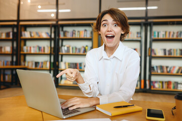 Wall Mural - Young surprised shocked happy successful employee IT business woman wearing white shirt casual clothes hold use work point finger on laptop pc computer sit at office desk. Achievement career concept.